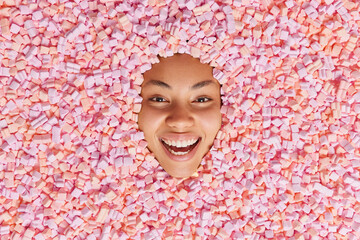 Happy smiling young ethnic woman sticks head in puffy appetizing marshmallow smiles toothily has...