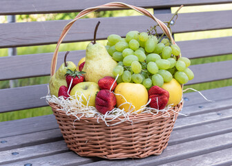 Round wicker basket full of fresh fruits and berries stands on a wooden bench
