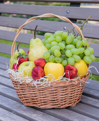 Round wicker basket full of fresh fruits and berries stands on a wooden bench