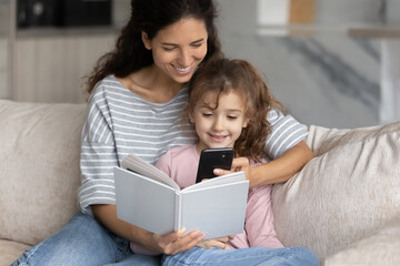 Happy Hispanic young mother and small daughter relax together read book talk on video call on cellphone. Smiling Latin mom and teen girl child rest at home enjoy story use modern smartphone.