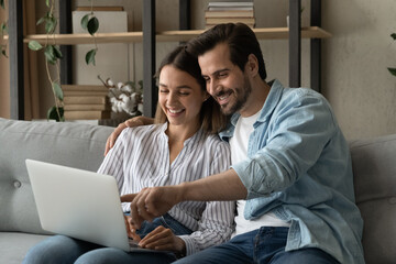 Happy beautiful couple sit on sofa enjoy easy purchasing on-line using laptop. Booking hotels,...
