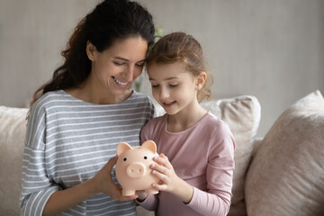Happy young Hispanic mom and small teen daughter feel provident economical make investment. Smiling Latino mother and girl child save money cash in piggybank. Banking, financial stability concept.