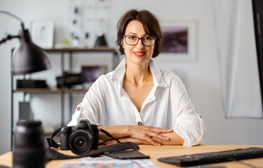 Portrait of adorable woman in white shirt and eyeglasses sitting at desk while working on computer. Female photographer posing at modern office.