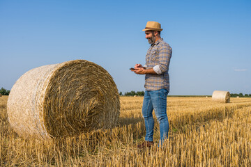 Naklejka na ściany i meble Happy farmer is standing beside bales of hay. He is examining straw after successful harvesting.