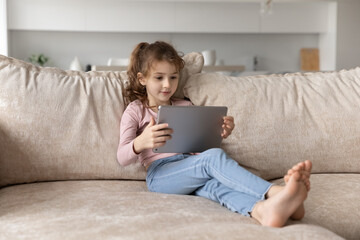 Smart small biracial girl child sit rest on sofa at home use modern tablet gadget playing. Happy ethnic kid relax on couch in living room talk speak on video webcam call on pad. Technology concept.