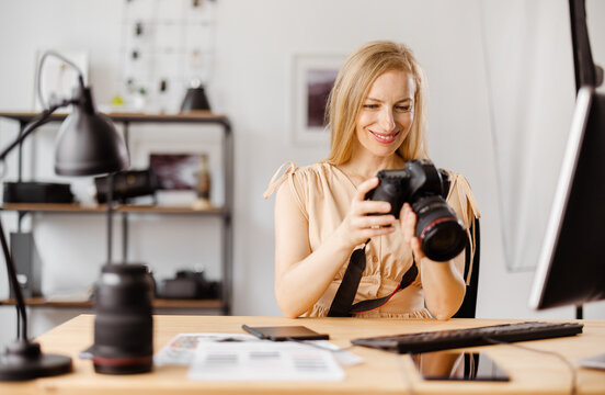 Beautiful woman with blond hair sitting at office and reviewing pictures on camera. Female photographer choosing photos for editing on computer.