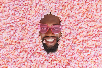 Positive bearded dark skinned guy sticks head in delicious puffy pink marshmallow enjoys eating...