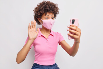 Afro American woman being on self isolation during coronavirus pandemic wears protective face mask makes video call waves palm in hello gesture keeps smartphone in front poses against white wall