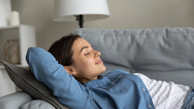 Close up beautiful serene carefree young woman relaxing on sofa, female put hands behind head closed eyes smile breath fresh air in modern living room. Stress-free day off, recreation, comfort concept