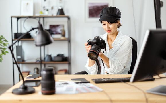Focused female photographer in stylish outfit reviewing pictures on digital camera while sitting at desk. Busy mature woman working at modern bright office.