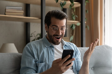 Young man in glasses sit on couch holds cellphone gawp at device screen with mouth open feels...