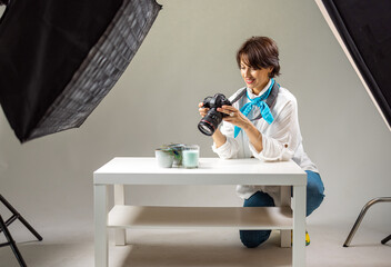Smiling woman squatting near white table and looking on screen of photo camera. Experienced photographer reviewing pictures taken at modern studio.