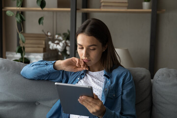 Young serious woman sit on sofa spend free time use modern wireless tech, read media news on tablet...