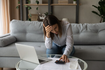 Desperate young woman sit on sofa at home calculates expenses feeling stressed about bank loan...