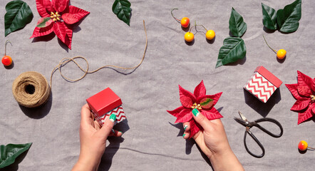 Christmas flat lay on natural beige linen textile. Hands decorate gift boxes. Red poinsettia, orange wild apple decor. Hemp cord, scissors, red and dark green poinsettia leaves .