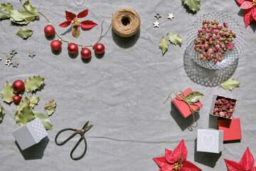 Christmas flat lay on natural beige linen textile. Gift boxes, red poinsettia, red baubles, winter decor with copy-space. Dry rose tea leaves. Hemp cord, scissors, red and dark green poinsettia leaves