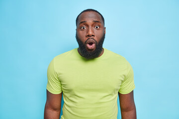 Portrait of dark skinned man looks embarrassed at camera has shocked expression keeps mouth opened wears casual green t shirt isolated over blue background gasps from fantastic news. Omg no way