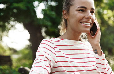 Portrait of happy smiling woman talking on mobile phone. Cute blond girl student sitting in park...
