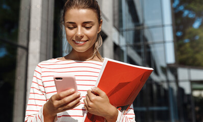 Portrait of smiling blond girl chatting on the way to universy or work. Woman holding books and looking at her mobile phone, message someone