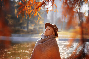 Cute young woman in scarf and hat enjoying breathing fresh air in autumn park with closed eyes