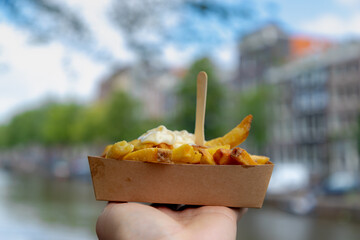 Selective focus of french fries in brown paper box on a man hand with blurred Amsterdam canal house, Friet or Patat served with mayonnaise and wooden cocktail fork, Dutch favorite snacks, Netherlands.