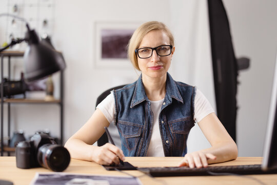 Female photographer in eyeglasses and casual clothes sitting office desk and editing photos on modern laptop. Concept of people and creative work.