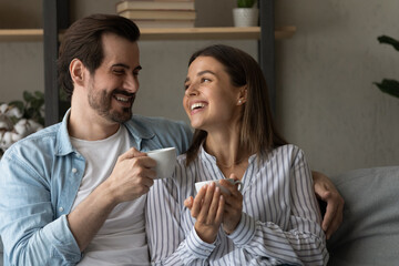Lovely 30s couple sit on sofa with tea cups enjoy conversation. Beautiful woman handsome man hugging resting together on sofa laughing joking having pleasant communication at home on carefree weekend