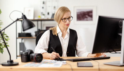 Focused photographer in eyeglasses sitting at workplace and retouching photos. Mature woman with...