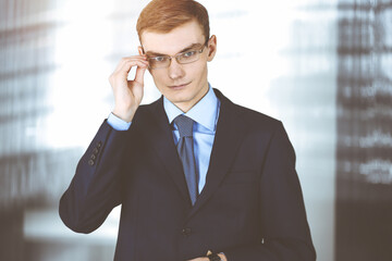 Young businessman wearing suit, with glasses, while standing in the sunny office. Business success concept nowadays