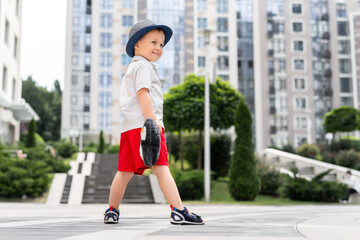 Cute adorable caucasian 3-4 years blond little kid boy in stylish shorts shirt and hat hold soft teddy toy enjoy playing outdoors at urban city center downtown area. Funny child portrait