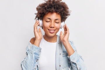 Portait of pleased Afro American woman enjoys good sound and favorite song keeps hands on wireless stereo headphones uses best free music app wears white t shirt and denim shirt poses indoor