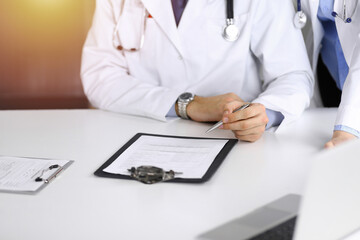Fototapeta premium Group of unknown doctors are sitting at the desk and discussing problems or disease treatment in sunny hospital office. Covid 2019