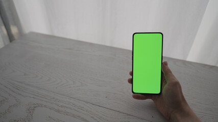 Man showing and using smarphone with green screen while sitting behind white oak table