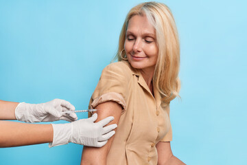 Injection antibody and immunization concept. Satisfied middle aged woman feels protected after making inoculation against coronavirus poses against blue background. Faceless doctor makes shot