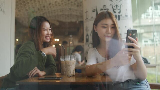 Asian woman friends sitting in cafe in the city and using smartphone taking selfie together. Female friendship enjoy and having fun outdoor lifestyle with wireless technology and modern gadget device