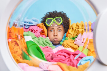 Funny dark skinned young woman with curly hair wears snorkeling mask on forehead blows cheeks poses around multicolored laundry blue background. Laundry worker loads washer. Appliance concept - obrazy, fototapety, plakaty