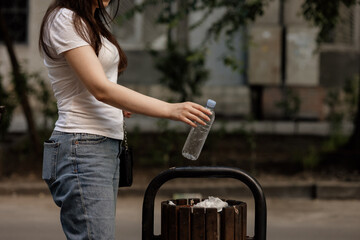 Closeup hand throwing plastic empty water bottle in recycling bin, isolated building and trees background. Hand of woman throwing plastic bottle into old trash can, concept of environmental 