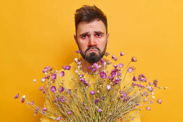 Spring allergy concept. Upset frustrated bearded man suffers from rhinitis holds bouquet of flowers...