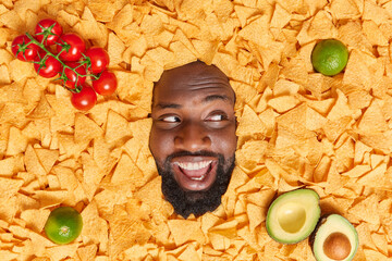 Positive dark skinned bearded man buried in nacho chips looks away with cheerful expression surrounded by red tomatoes lime and avocado going to eat tasty snack. Overhead view. Food concept.