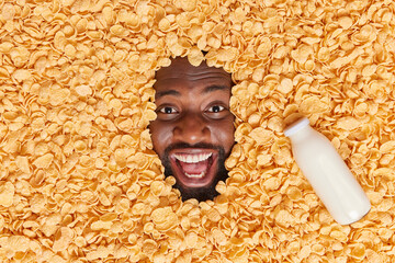 Eating healthy food. Positive dark skinned man buried in heap of cornflakes with bottle of milk near keeps mouth wide opened has happy mood expresses positive emotions. Ingredients for breakfast