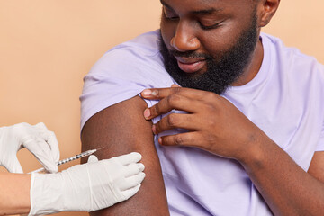 Nurse in white medical gloves makes vaccination in shoulder of patient. Dark skinned bearded adult...