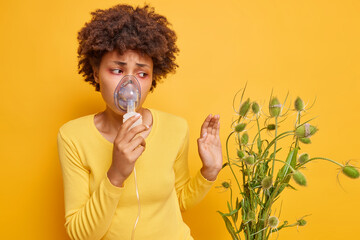 Unhappy dark skinned woman suffers from allergy to wild flowers uses inhalation nebulizer has red...