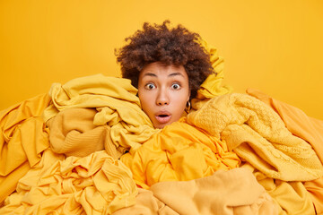 Stunned curly haired ethnic woman with stuck head in stack of clothes buys different items of...