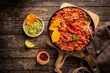 Mexican hot chili con carne in a pan with tortilla chips on dark background, top view