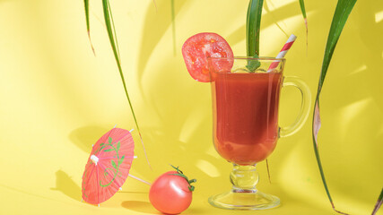 Glass glass with tomato juice in the shade of leaves on a yellow background.