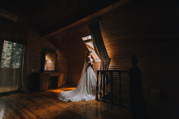 A beautiful bride in a white long dress stands near a window and a mirror in a wooden room made of logs. Wedding portrait.