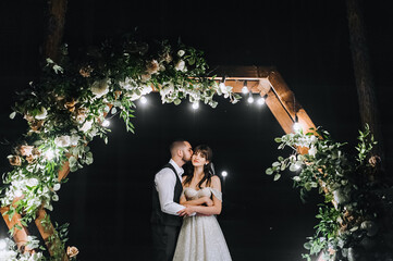 A young groom in a vest and a beautiful bride in a white long dress tenderly embrace, kiss near a...