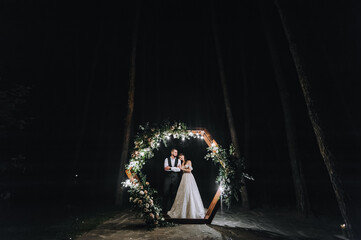 Stylish groom in a vest and a cute brunette bride in a white long dress with a veil tenderly hug...