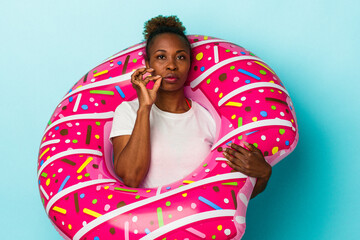 Young african american woman with inflatable donut isolated on blue background with fingers on lips keeping a secret.