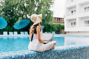 Fototapeta premium At a resort hotel near the pool sits a young happy woman in a straw hat with a laptop. technology concept and remote work.